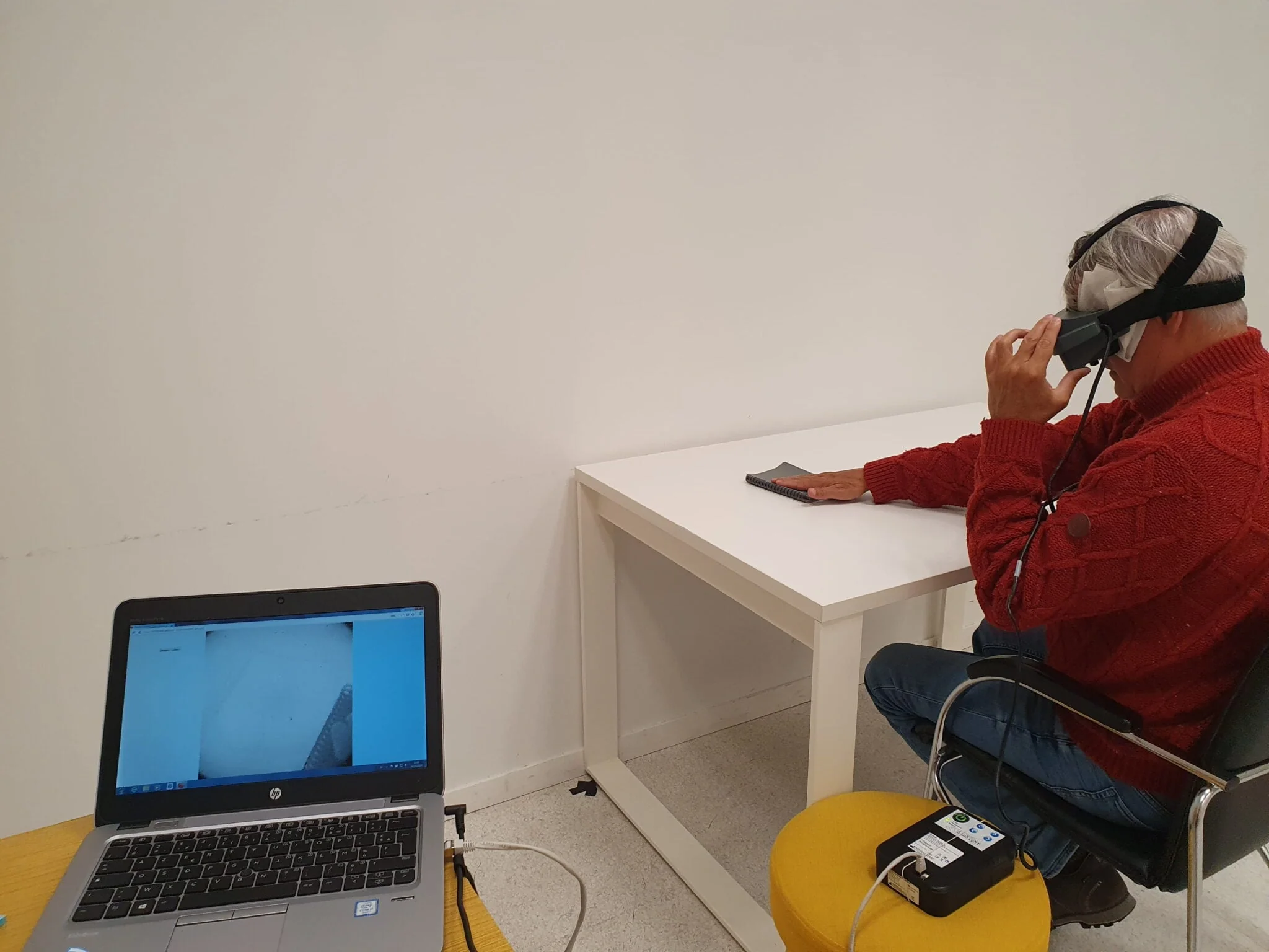 Participant assis portant des lunettes spéciales et touchant un carnet sur une table blanche, avec un ordinateur à proximité.